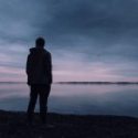 Silhouette of a person standing at dusk near a body of water. The sky is a mix of blues and pinks, reflecting on the water, creating a serene and contemplative mood.